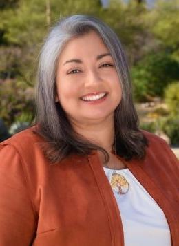 portrait of Erika Quiñonez wearing a rust jacket and white blouse posing in front of greenery. 